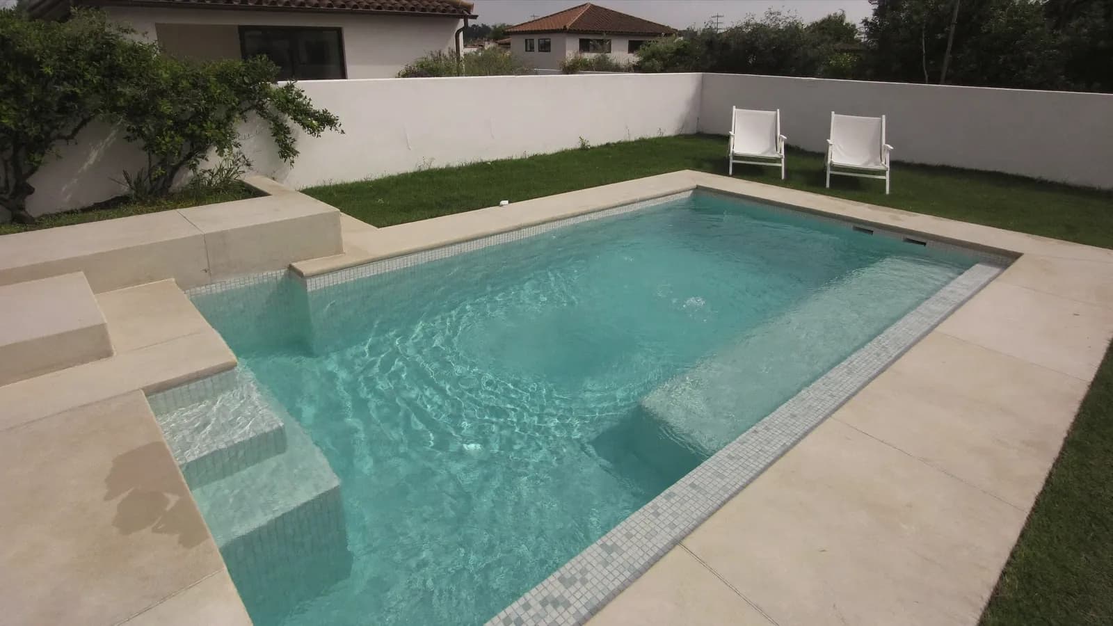 Piscina rectangular de hormigón recién terminada en un jardín, con gresite blanco que da al agua un color turquesa, coronación de piedra clara y escalera de obra en una esquina