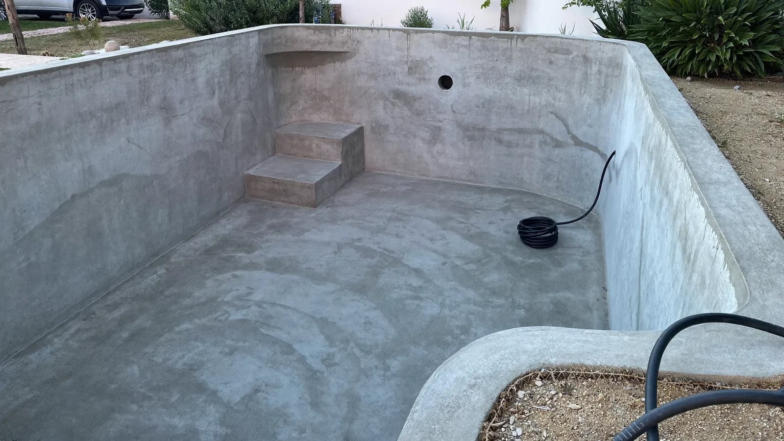 Vaso de piscina recién proyectado con hormigón, todavía húmedo, con escalera de obra en una esquina y una manguera enrollada en el fondo